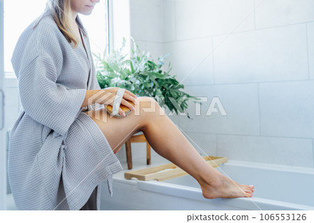 Young woman in bathrobe doing scrubbing massage with dry brush before morning shower in light colored bathroom. Anti-cellulite treatment, lymphatic drainage massage. Women beauty. Selective focus 106553126