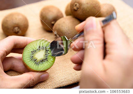 A man's hand holding a sliced kiwi and eating it with a spoon. A heap of kiwi fruit on a jute bag 106553683