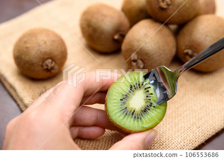 A man's hand holding a sliced kiwi and eating it with a spoon. A heap of kiwi fruit on a jute bag 106553686