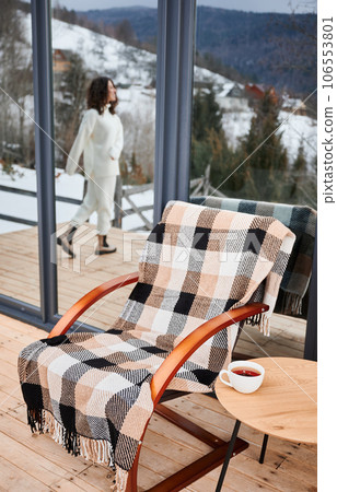Chair and table with cup of tea on on terrace of modern barn house in winter mountains. Happy woman tourist reflected in huge window. Chair and table with cup of tea on on terrace of modern barn house in winter mountains. Happy woman tourist reflected in huge window. 106553801