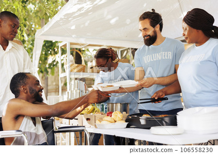 Volunteers from varying ethnicities come together with smiles offering assistance to handicapped poor and homeless. Charity group provided free food to black man in wheelchair meeting his needs. 106558230