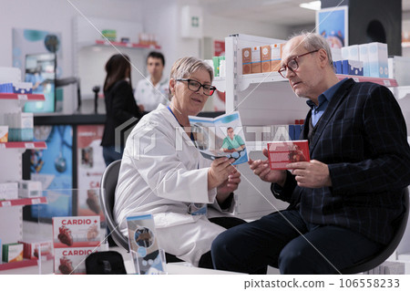 Pharmacy worker talking to elderly customer about heart medications and showing medical healthcare brochure. Professional drugstore assistant helping senior man choosing supplements Pharmacy worker talking to elderly customer about heart medications and showing medical healthcare brochure. Professional drugstore assistant helping senior man choosing supplements 106558233