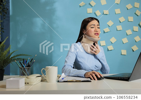 Asian woman with cervical collar on neck using laptop computer feeling discomfort and pain. Female corporate employee with injury working seated at startup company workstation. Asian woman with cervical collar on neck using laptop computer feeling discomfort and pain. Female corporate employee with injury working seated at startup company workstation. 106558234