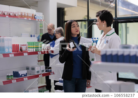 Pharmaceutical assistant and young asian woman discussing vitamin complex components in drugstore. Pharmacy employee giving assistance to customer in choosing multivitamin capsules Pharmaceutical assistant and young asian woman discussing vitamin complex components in drugstore. Pharmacy employee giving assistance to customer in choosing multivitamin capsules 106558237