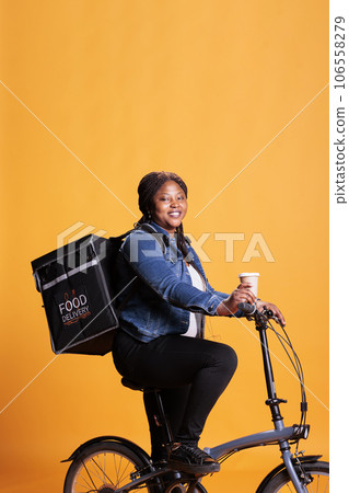 Portrait of restaurant courier courier holding cup of coffee while delivering takeout food order to customers using bike as transportation. Pizzeria worker standing in studio with yellow background. Portrait of restaurant courier courier holding cup of coffee while delivering takeout food order to customers using bike as transportation. Pizzeria worker standing in studio with yellow background. 106558279