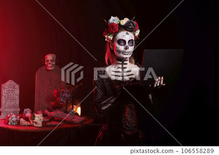 Model with halloween costume holding laptop, posing on tranditional mexican holiday celebration. Wearing skull make up and day of the dead costume with body art, goddess of death. Model with halloween costume holding laptop, posing on tranditional mexican holiday celebration. Wearing skull make up and day of the dead costume with body art, goddess of death. 106558289