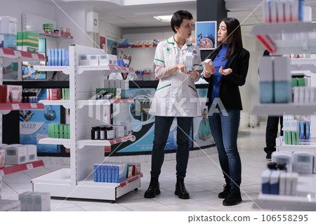 Drugstore consultant helping woman customer choosing sunscreen lotion, explaining spf numbers differences. Pharmaceutical specialist and young asian woman talking about skin care products Drugstore consultant helping woman customer choosing sunscreen lotion, explaining spf numbers differences. Pharmaceutical specialist and young asian woman talking about skin care products 106558295