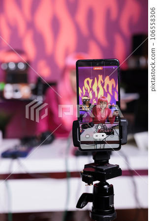 Influencer filming podcast using mobile phone camera discussing lifestyle advice with subscribers, posting content on her channel. Asian vlogger standing in broadcast studio recording vlog Influencer filming podcast using mobile phone camera discussing lifestyle advice with subscribers, posting content on her channel. Asian vlogger standing in broadcast studio recording vlog 106558306