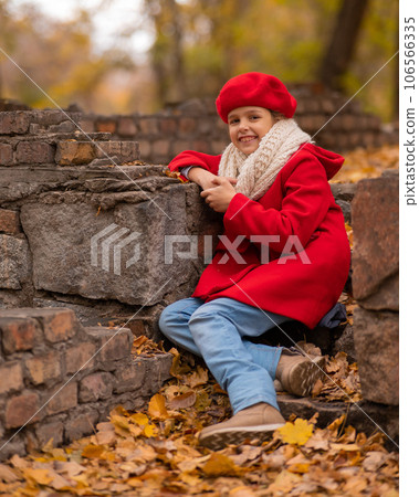 Smiling caucasian girl in a red coat and beret sits on a brick wall on a walk in autumn. Smiling caucasian girl in a red coat and beret sits on a brick wall on a walk in autumn. 106566335