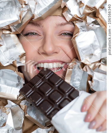 The face of a Caucasian woman surrounded by candy wrappers. The girl eats a bar of chocolate. 106566340