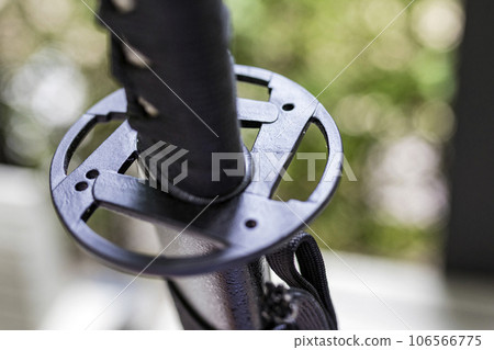 A shallow focus shot of Tsuba, or hand guard, on the handle of a katana (Japanese long sword) It is a work of art that is widely popular and has a high price. The background is a blurred fence line. A shallow focus shot of Tsuba, or hand guard, on the handle of a katana (Japanese long sword) It is a work of art that is widely popular and has a high price. The background is a blurred fence line. 106566775