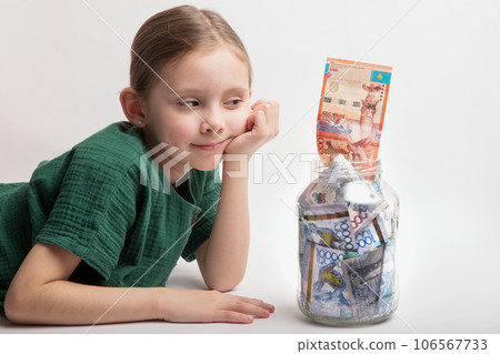 Kid with a large transparent jar with Kazakhstani tenge money on a white background Kid with a large transparent jar with Kazakhstani tenge money on a white background 106567733