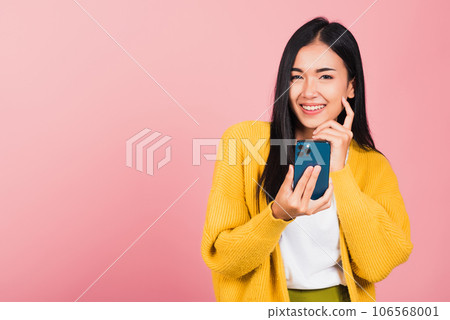 Happy Asian portrait beautiful cute young woman excited smiling holding mobile phone and think idea finger touch face, studio shot isolated on pink background, female using smartphone making gesture 106568001