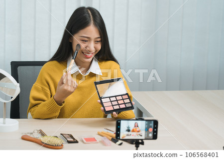 Young asian woman vlogging about cosmetics skin care items products on table with her video camera and demonstrates product use and reviews for her online blog channel. 106568401