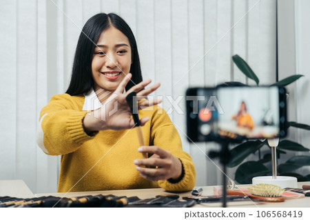 Young asian woman vlogging about cosmetics skin care items products on table with her video camera and demonstrates product use and reviews for her online blog channel. 106568419