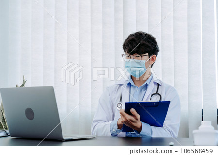 Doctor was sitting in the office working on a laptop computer while wearing a mask. 106568459