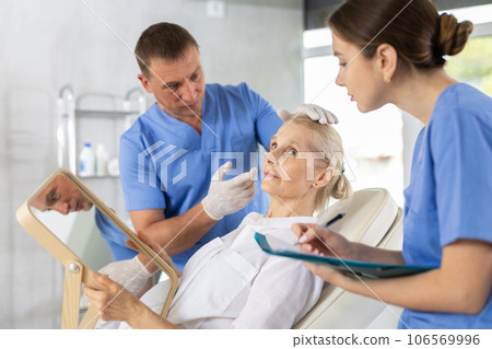 Cosmetologist with assistant explaining facial procedure to elderly woman 106569996