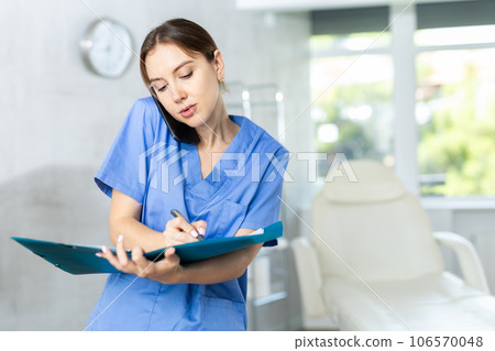 Nurse holding mobile phone between ear and shoulder talking and writing notes to clipboard 106570048