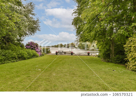 a park with trees and flowers in the foregrounds, taken from behind it is an empty bench on the grass 106570321