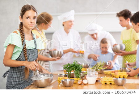 Cooking course - portrait of a girl in an apron who is learning to beat eggs to make pancakes at cooking class Cooking course - portrait of a girl in an apron who is learning to beat eggs to make pancakes at cooking class 106570474