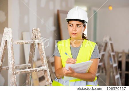 Woman foreman in a protective helmet and yellow vest checks the completed construction work on drawing 106570537