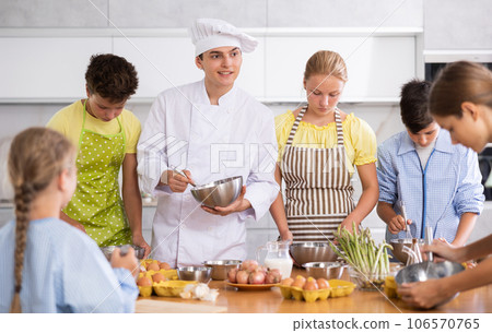 Group of teenagers and friendly chef cook cooking in modern kitchen. Learn to beat raw eggs Group of teenagers and friendly chef cook cooking in modern kitchen. Learn to beat raw eggs 106570765