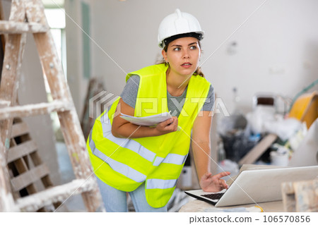 Female foreman in a protective helmet and a yellow vest checks the execution of repair work using laptop 106570856