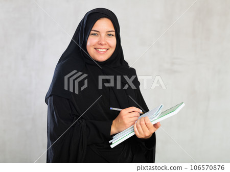 gleeful girl in hijab holds several notebooks and writes important note in her diary gleeful girl in hijab holds several notebooks and writes important note in her diary 106570876