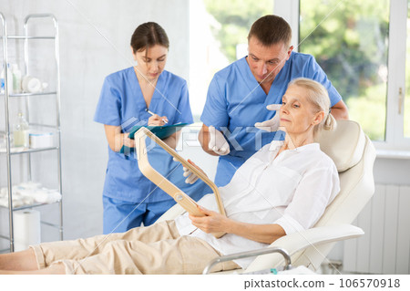 Man dermatologist examines skin of senior womans face, female nurse records data in client card 106570918