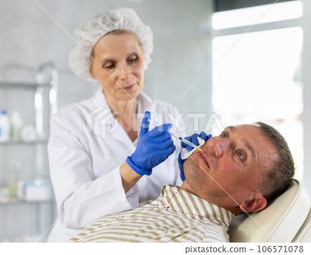 Mature woman doctor gives injection and performs procedure of lip augmentation of male patient 106571078