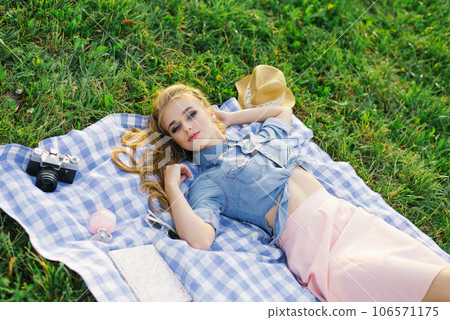 Stylish young woman with bright makeup in a denim shirt is lying on her back on a plaid in the open air and resting Stylish young woman with bright makeup in a denim shirt is lying on her back on a plaid in the open air and resting 106571175