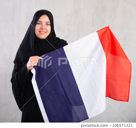 Balanced smiling Muslim woman in traditional black hijab holds flag of France Balanced smiling Muslim woman in traditional black hijab holds flag of France 106571321