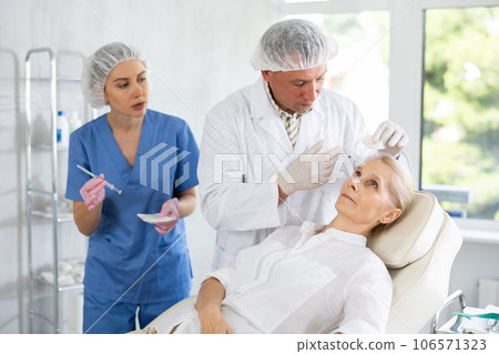 Professional cosmetologist doing injections in forehead to elderly woman Professional cosmetologist doing injections in forehead to elderly woman 106571323