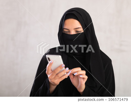 Young woman dressed in traditional islamic clothes with her face covered looking at her phone Young woman dressed in traditional islamic clothes with her face covered looking at her phone 106571479
