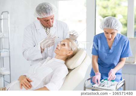 Professional cosmetologist doing injections in forehead to elderly woman 106571480