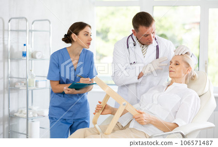 Man dermatologist examines skin of senior womans face, female nurse records data in client card Man dermatologist examines skin of senior womans face, female nurse records data in client card 106571487