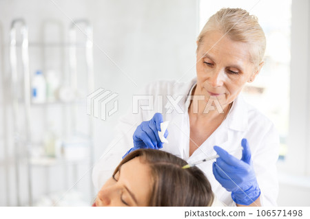 Old woman doctor implementing hair care procedure by injection method 106571498