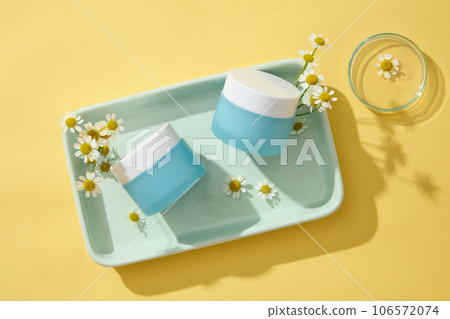 Beauty blue jars inside a dish decorated on yellow background with Chamomilla flowers. Chamomilla (Matricaria chamomilla) helping to arm our skin against environmental damage 106572074