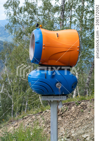 Yellow snow cannon on the edge of a hill overlooking a valley in summer or warm winter, green mountain peaks. Ski slope without snow in summer or during warm winter, greenhouse effect concept. Yellow snow cannon on the edge of a hill overlooking a valley in summer or warm winter, green mountain peaks. Ski slope without snow in summer or during warm winter, greenhouse effect concept. 106572092
