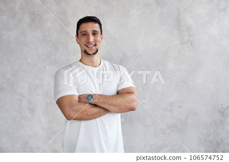 Brunette man smiles, looks at camera, crossed arms over chest on white textured wall background, natural light 106574752