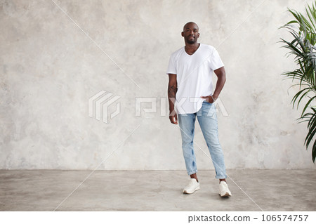 Handsome African American young man stands full body, looks at camera, against bright textured wall in studio. Male model wears casual clothes white T-shirt and jeans 106574757