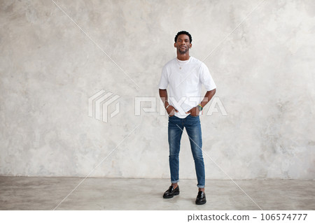 Full length African American young man stands against bright textured wall in studio. Male model keeps hands in pockets and wears casual clothes white T-shirt, jeans Full length African American young man stands against bright textured wall in studio. Male model keeps hands in pockets and wears casual clothes white T-shirt, jeans 106574777