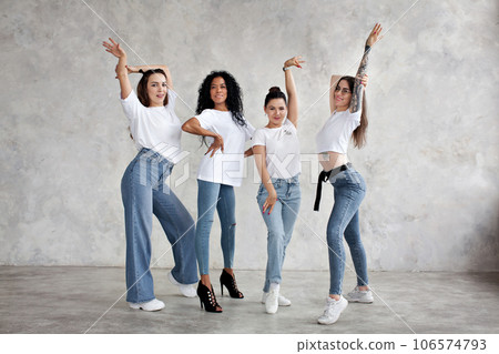 Smiling multiethnic women pose, dance, raise hands up against gray background of studio. Group of girls are similar dressed in jeans and white T-shirts 106574793