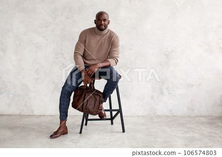 Stylish man sitting on chair and looking at camera. Confident fashion model in fashionable outfit turtleneck knitted sweater, plaid trousers, leather shoes and bag. Office clothes style for busines 106574803