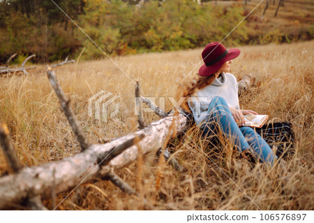 Fall picnic in nature. Young woman sitting on the blanket reading a book and smiling outdoors. Fall picnic in nature. Young woman sitting on the blanket reading a book and smiling outdoors. 106576897