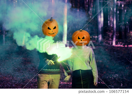 A couple with Halloween pumpkin on head in a woodland night with colourful smoke. Halloween Jack-o-lantern funny party. A couple with Halloween pumpkin on head in a woodland night with colourful smoke. Halloween Jack-o-lantern funny party. 106577154