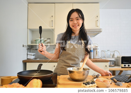 Cheerful asian housewife with spatula in hand, preparing breakfast in kitchen. People, food and domestic life concept 106579885