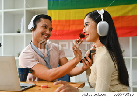 Cheerful gay man applying powder with brush on face female friend. LGBT, friendship and lifestyle concept 106579889