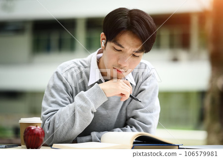Concentrated asian male student reading book, preparing for exam or, doing homework remotely in the park Concentrated asian male student reading book, preparing for exam or, doing homework remotely in the park 106579981