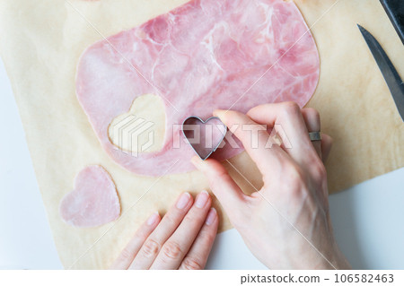 The cook cuts heart-shaped sausage-ham cut into heart shape on parchment, top view, close-up. Cooking pizza for Valentine's Day. 106582463